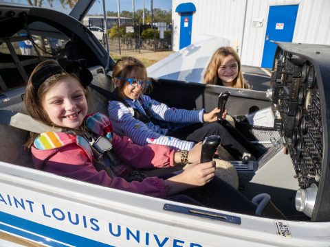 Two happy young girls sitting in the cockpit of a plane with a friend standing nearby