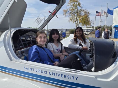 Two young women sitting in plane cockpit with canopy open, with another woman standing next to the plane
